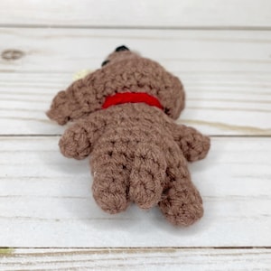 May include: A handmade brown crochet dog toy with a red collar. The dog has black eyes and a small white ear. The toy is lying on its side, showcasing its crocheted texture and shape. A charming, handcrafted item.