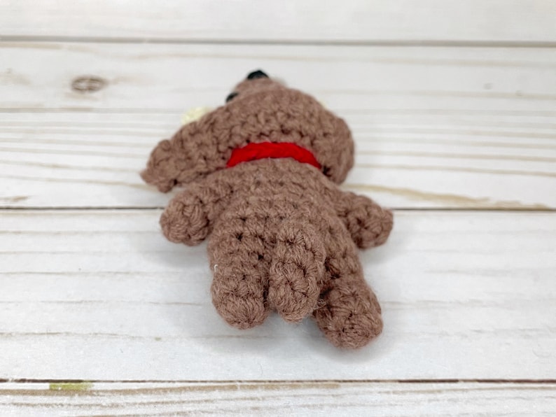 May include: A handmade brown crochet dog toy with a red collar. The dog has black eyes and a small white ear. The toy is lying on its side, showcasing its crocheted texture and shape. A charming, handcrafted item.