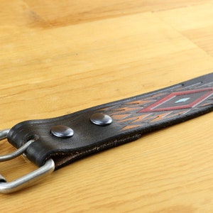 May include: A black leather belt with a geometric pattern in shades of orange, red, and blue. The belt has a silver buckle and two silver rivets.