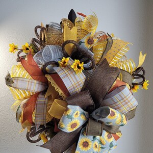 May include: A decorative wreath featuring a variety of ribbons in shades of brown, yellow, orange, and gray. The wreath includes ribbons with sunflower patterns, plaid designs, and solid colors. Small artificial sunflowers are also incorporated into the design.