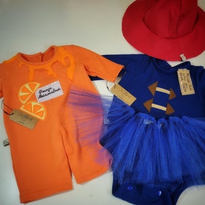 Puede incluir: Dos conjuntos de bebé: un mono naranja con aplicaciones de rodajas de naranja y un mono azul con un diseño de estrella marrón. Ambos conjuntos tienen faldas de tul azules a juego. También se incluye un sombrero rojo.