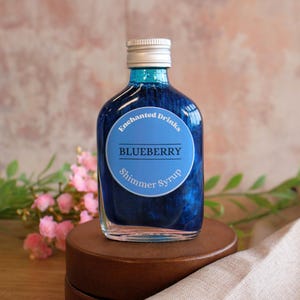 May include: A clear glass bottle filled with deep blue liquid containing shimmering particles. The bottle has a silver screw cap and a round blue label with the text Enchanted Drinks BLUEBERRY Shimmer Syrup printed in white and dark blue.