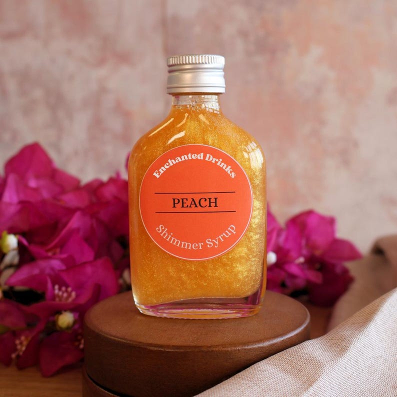 May include: A clear glass bottle filled with gold-colored shimmer syrup. The bottle has a silver screw cap and a round orange label with the text Enchanted Drinks, PEACH, and Shimmer Syrup printed in white and black lettering.