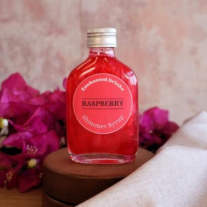May include: A clear glass bottle filled with vibrant red raspberry shimmer syrup. The bottle features a round label with the text Enchanted Drinks, RASPBERRY, and Shimmer Syrup in white and black lettering. It has a silver screw-top cap.
