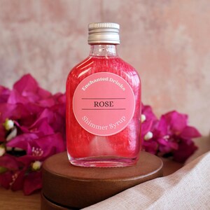 May include: A clear glass bottle filled with vibrant pink shimmer syrup, topped with a silver screw cap. A round label on the front features the text Enchanted Drinks, ROSE, and Shimmer Syrup in white and black lettering against a pink background.