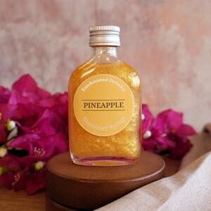 May include: A clear glass bottle filled with golden pineapple shimmer syrup. The bottle has a silver screw cap and a round yellow label that reads Enchanted Drinks PINEAPPLE Shimmer Syrup in white and black text.