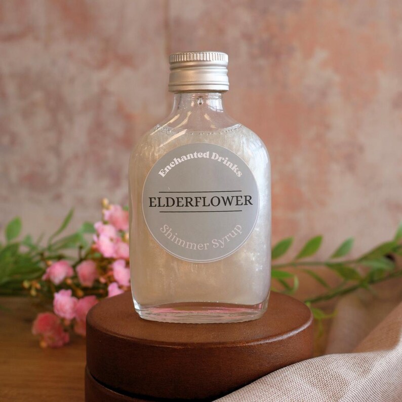 May include: A clear glass bottle filled with a pearlescent liquid, topped with a silver screw cap. The circular label reads Enchanted Drinks ELDERFLOWER Shimmer Syrup in white and black text. The bottle has a flat, rounded rectangular shape.