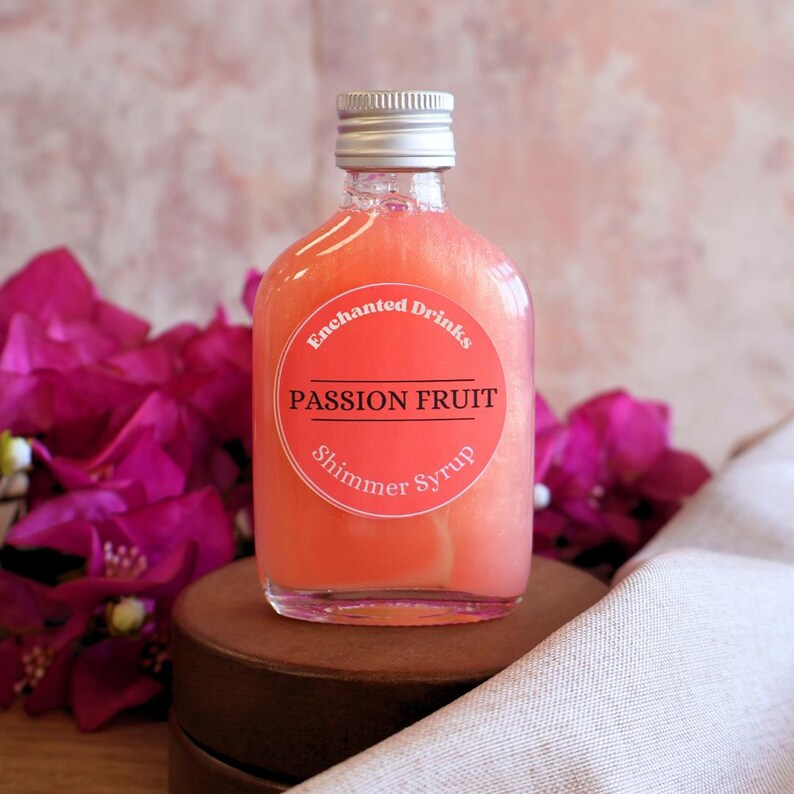 May include: A clear glass bottle filled with a pink, pearlescent passion fruit shimmer syrup. The bottle has a silver screw cap and a round orange label that reads Enchanted Drinks, PASSION FRUIT, and Shimmer Syrup in white and black text.