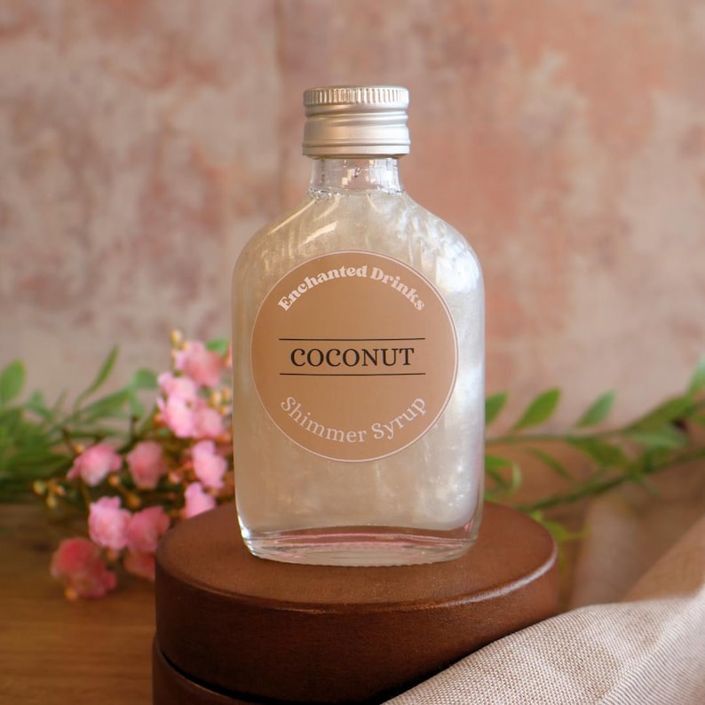May include: A clear glass bottle filled with a pearlescent white liquid, topped with a silver screw cap. The round label features the text Enchanted Drinks, COCONUT, and Shimmer Syrup in dark brown lettering against a tan background.