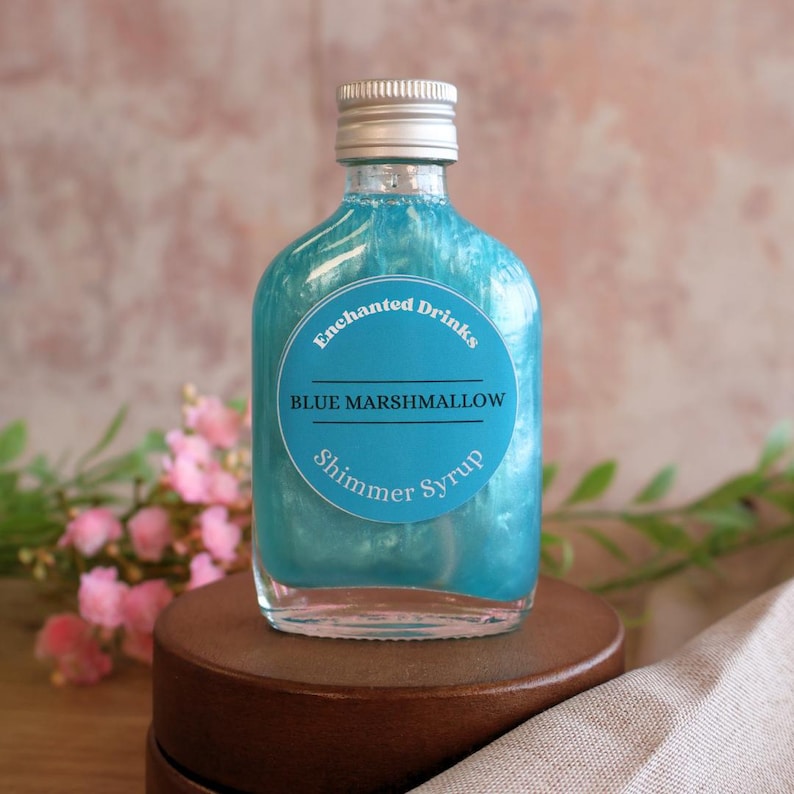 May include: A clear glass bottle filled with a vibrant blue liquid that has a pearlescent shimmer effect. The bottle features a round blue label with the text Enchanted Drinks, BLUE MARSHMALLOW, and Shimmer Syrup, topped with a silver screw cap.