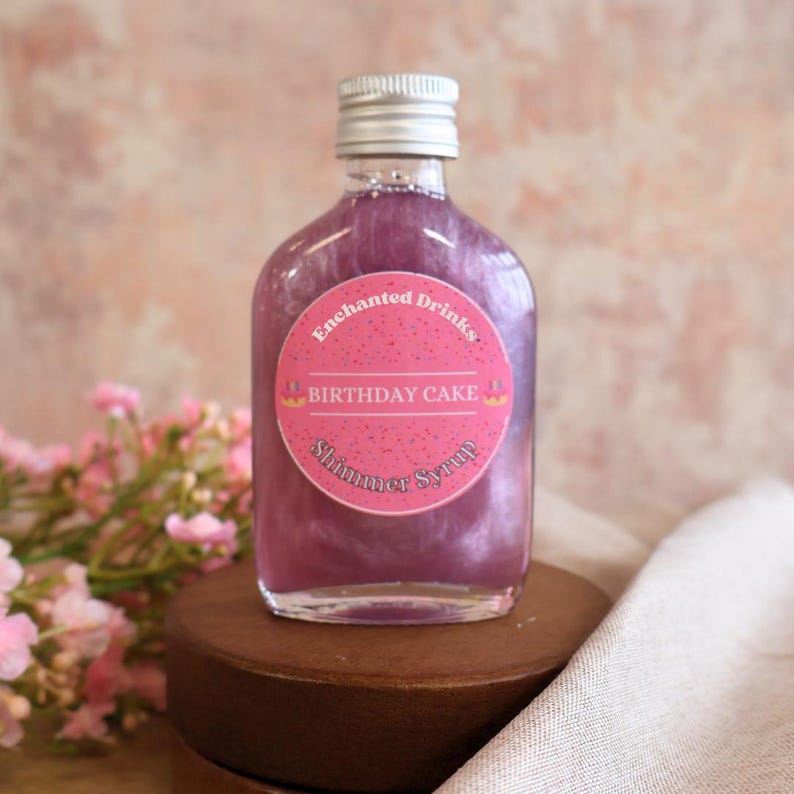 May include: A clear glass bottle filled with purple shimmer syrup, topped with a silver screw cap. A round pink label on the front features the text Enchanted Drinks, BIRTHDAY CAKE, and Shimmer Syrup, decorated with small cupcake icons.