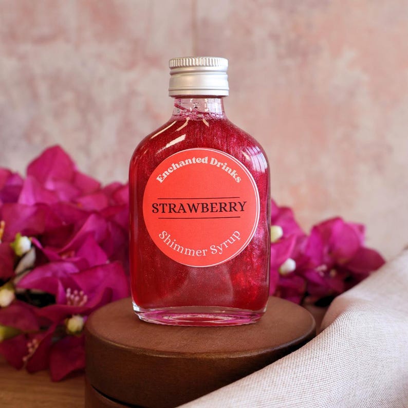 May include: A clear glass bottle filled with a vibrant red strawberry shimmer syrup. The bottle has a silver screw cap and a round label that reads Enchanted Drinks STRAWBERRY Shimmer Syrup in black and white text against a red background.