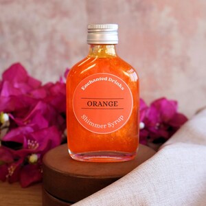 May include: A clear glass bottle filled with vibrant orange shimmer syrup. The bottle has a silver screw-top cap and a round orange label that reads Enchanted Drinks, ORANGE, and Shimmer Syrup in white and black text.