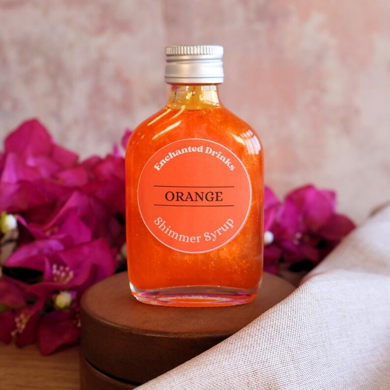 May include: A clear glass bottle filled with vibrant orange shimmer syrup. The bottle has a silver screw-top cap and a round orange label that reads Enchanted Drinks, ORANGE, and Shimmer Syrup in white and black text.