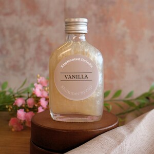 May include: A clear glass bottle filled with a shimmering gold liquid, topped with a silver screw cap. A circular label on the front reads Enchanted Drinks, VANILLA, and Shimmer Syrup in black and white text against a light beige background.