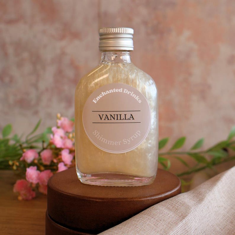 May include: A clear glass bottle filled with a shimmering gold liquid, topped with a silver screw cap. A circular label on the front reads Enchanted Drinks, VANILLA, and Shimmer Syrup in black and white text against a light beige background.