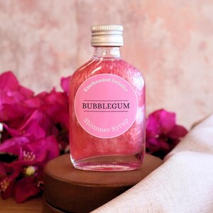 May include: A clear glass bottle filled with pink shimmer syrup. The bottle has a silver screw cap and a round pink label that reads Enchanted Drinks BUBBLEGUM Shimmer Syrup in white and black text. The liquid inside has a swirling pearlescent effect.
