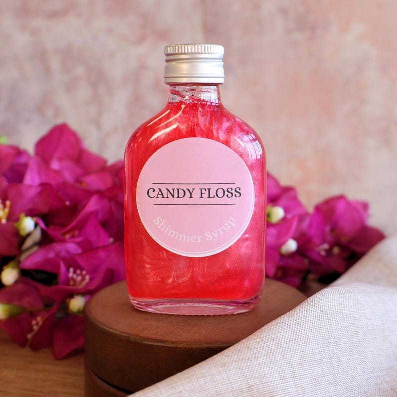 May include: A clear glass bottle filled with vibrant pink shimmer syrup, topped with a silver screw cap. A circular white label on the front features the black text CANDY FLOSS and the smaller text Shimmer Syrup in a light pink font.