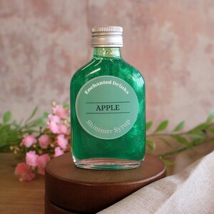 May include: A clear glass bottle filled with vibrant green shimmer syrup. The bottle features a round label with the text Enchanted Drinks, APPLE, and Shimmer Syrup. It has a silver screw-top cap and a rounded, flat-bottomed flask shape.