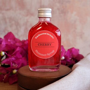 May include: A clear glass bottle filled with vibrant red shimmer syrup. The bottle has a silver screw-top lid and a round red label that reads Enchanted Drinks, CHERRY, and Shimmer Syrup in white and black text. The liquid has a metallic sheen.