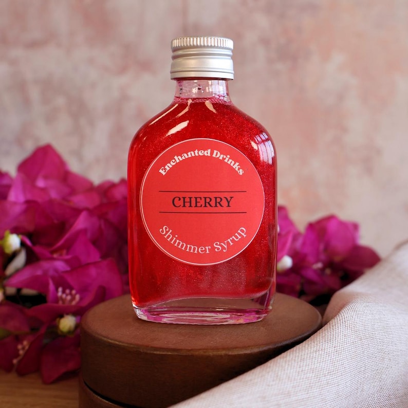 May include: A clear glass bottle filled with vibrant red shimmer syrup. The bottle has a silver screw-top lid and a round red label that reads Enchanted Drinks, CHERRY, and Shimmer Syrup in white and black text. The liquid has a metallic sheen.