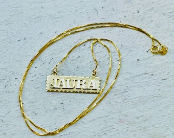 May include: A gold chain necklace with a rectangular pendant that says "LAURA" in block letters.