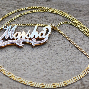 May include: A gold chain necklace with a nameplate pendant that says "Marsha" in a cursive font. The nameplate is two-toned, with a gold outline and a white center.