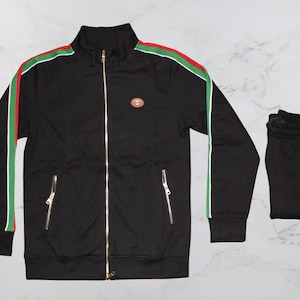 May include: Black track suit set with a jacket and matching pants. The jacket has a stand-up collar, gold zippers, and a red, white, and green stripe detail on the sleeves. The pants are folded and have gold zippers.