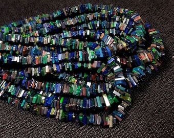 May include: A strand of multi-colored opal gemstone beads. The beads are rectangular in shape and have a variety of colors, including blue, green, black, and red. The beads are arranged in a loose pile on a dark gray fabric background.