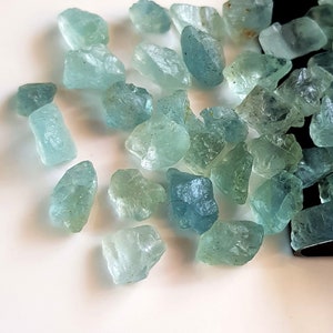May include: A collection of light blue aquamarine gemstones of various sizes and shapes. The gemstones are scattered on a white surface.