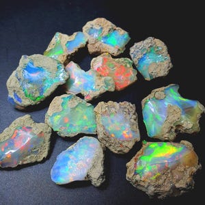 May include: A collection of rough opal stones, showcasing vibrant internal colors. The opals display a spectrum of hues, including blues, greens, and reds, set against a backdrop of natural, earthy matrix. The stones vary in size and shape, with a rough, unpolished texture.