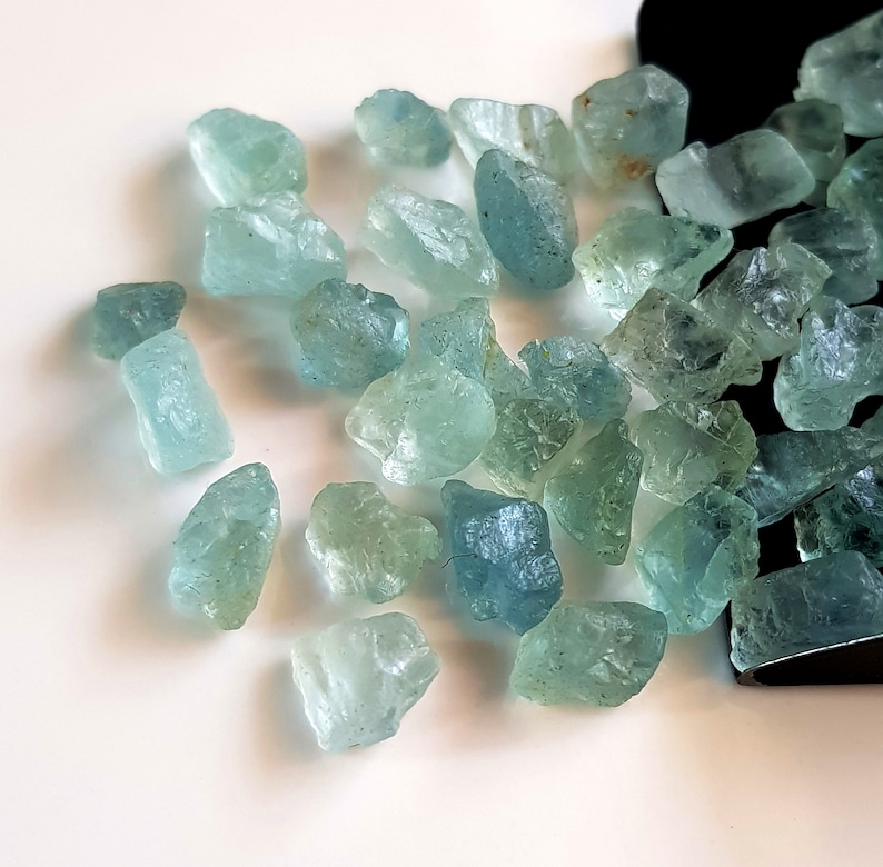 May include: A collection of light blue aquamarine rough gemstones. The stones are various sizes and shapes, with some having a more translucent appearance than others.