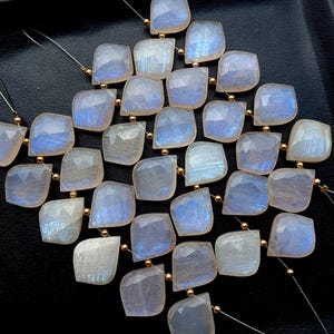 May include: A collection of square-cut moonstone beads with a pearlescent sheen and flashes of blue. Each bead is strung with gold-colored accents, creating a shimmering effect. The beads are arranged on a dark surface.