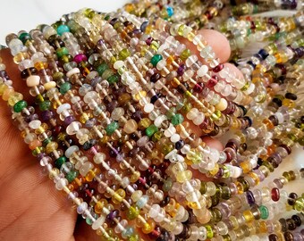 May include: A handful of colorful gemstone beads in various shapes and sizes. The beads are strung together, displaying a mix of colors including green, yellow, purple, and clear. The beads are held in a hand, showcasing the variety of the gemstones.