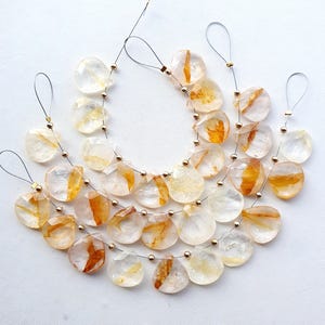 May include: Strands of clear, teardrop-shaped beads with orange and yellow inclusions. Each bead is strung on a silver wire with a small gold bead. The beads are arranged on a white surface.