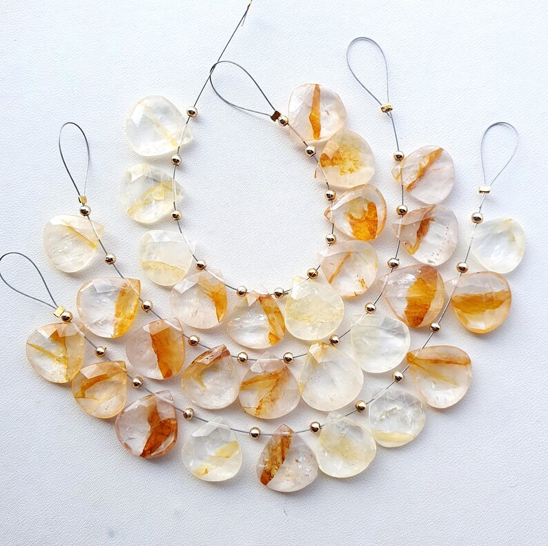 May include: Strands of clear, teardrop-shaped beads with orange and yellow inclusions. Each bead is strung on a silver wire with a small gold bead. The beads are arranged on a white surface.
