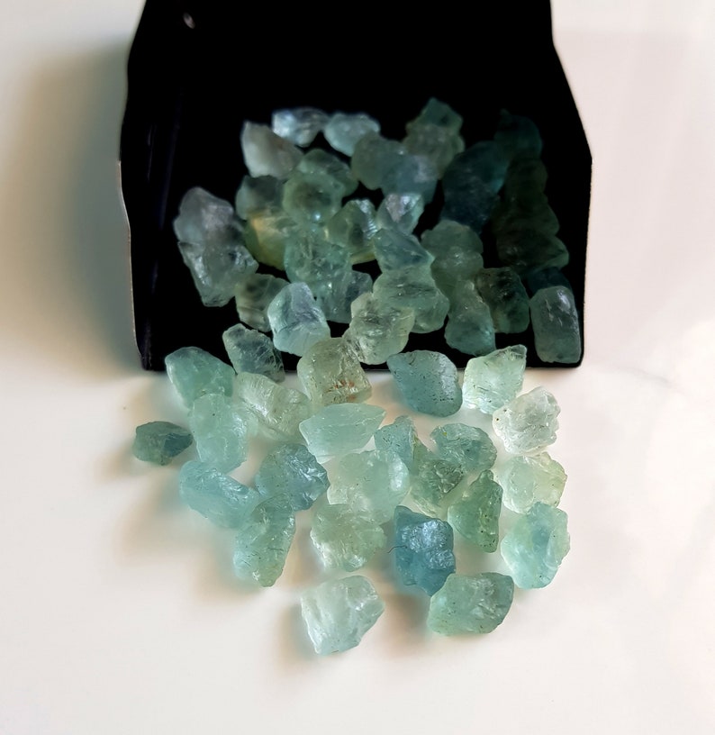 May include: A pile of light blue and green aquamarine rough gemstones. The gemstones are various sizes and shapes.