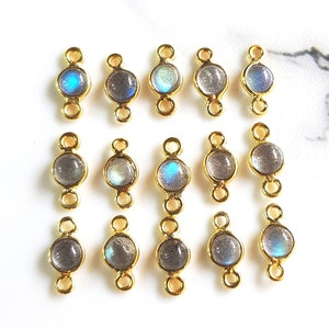 May include: A collection of fourteen gold-toned metal jewelry connectors, each featuring a circular labradorite gemstone. The stones display iridescent blue and grey flashes within a smooth bezel setting, with small loops at the top and bottom.