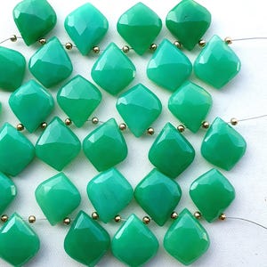 Könnte beinhalten: Eine Sammlung hellgrüner, facettierter Edelsteinperlen. Jede Perle hat eine rautenförmige Form mit mehreren Facetten und ist mit goldfarbenen Perlen aufgefädelt. Die Edelsteine haben eine polierte, reflektierende Oberfläche und sind in einem Raster angeordnet.