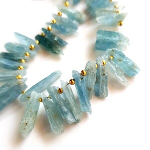 May include: A strand of light blue aquamarine gemstone beads with gold spacers. The beads are long and slender, with a slightly irregular shape.
