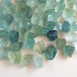 May include: A collection of aquamarine gemstones, ranging in shades of blue and green, with some having a more opaque appearance. The stones are rough and unpolished, with irregular shapes and sizes.