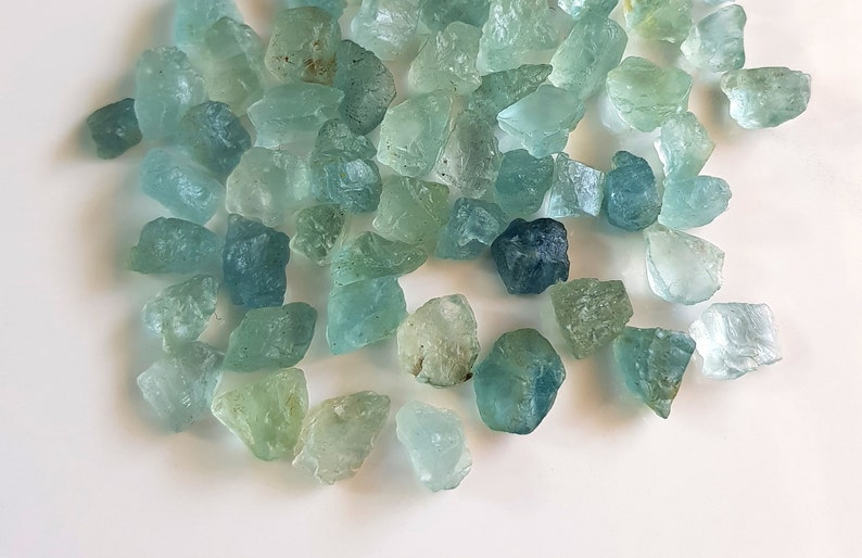May include: A collection of aquamarine gemstones, ranging in shades of blue and green, with some having a more opaque appearance. The stones are rough and unpolished, with irregular shapes and sizes.