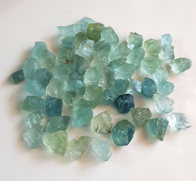 May include: A collection of rough aquamarine gemstones in various shades of blue and green. The stones are irregular in shape and have a natural, unpolished finish.