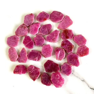 May include: A collection of raw, uncut ruby gemstones. The stones vary in size and shape, displaying a range of pink and deep red hues. The rough texture of the gems is visible, with natural imperfections. The rubies are scattered on a white surface.