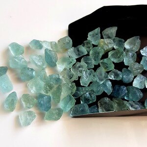 May include: A collection of light blue aquamarine rough gemstones. The gemstones are various sizes and shapes, and some have a slightly darker blue hue.