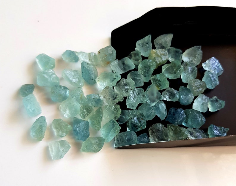 May include: A collection of light blue aquamarine rough gemstones. The gemstones are various sizes and shapes, and some have a slightly darker blue hue.