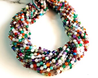 May include: A strand of faceted gemstone beads in a variety of colors, including red, green, purple, and orange. The beads are strung together, creating a colorful and textured appearance. The strand is on a white background.