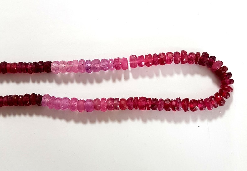 May include: A strand of pink and red gemstone beads. The beads are faceted and have a slightly irregular shape. The beads are strung on a clear thread.