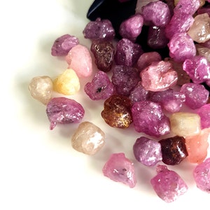 May include: A close-up of a pile of pink, red, and yellow rough gemstones. The gemstones are various sizes and shapes, and they are scattered on a white surface.