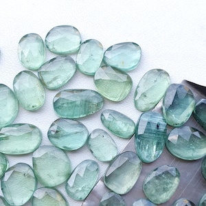 May include: A collection of light green, faceted, oval-shaped gemstones. The stones are scattered on a white surface.